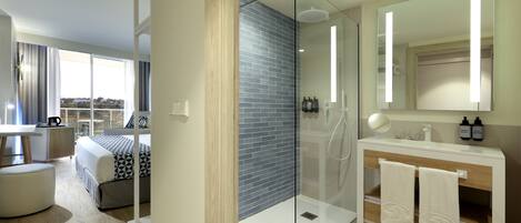 Une chambre d’hôtel moderne dotée d’une douche en verre, d’un grand lit avec une couette à motifs géométriques, d’un plancher en bois, d’une coiffeuse blanche avec un miroir et d’une vue sur l’extérieur grâce à une grande fenêtre.