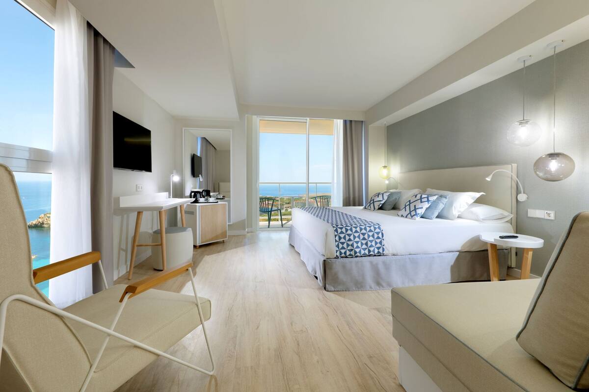 A modern hotel room with a large bed, a desk, a chair, and a view of the sea.