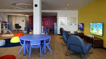 Un área de juegos infantil moderna con una mesa y sillas azules, estructuras de juego coloridas y una gran pantalla de televisión.