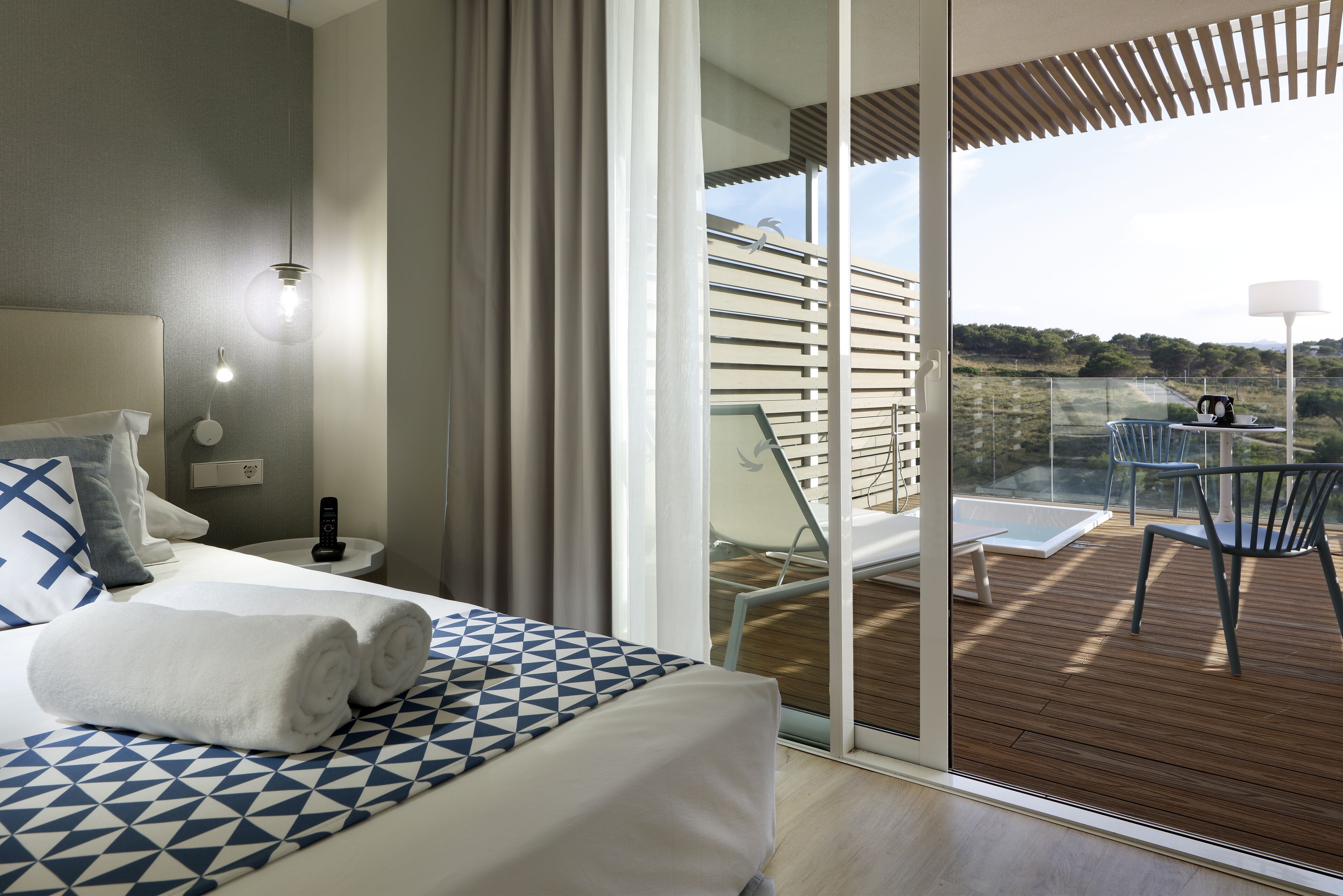A modern hotel room with a large bed, a bedside table, a nightstand, and a view of the outdoor deck.
