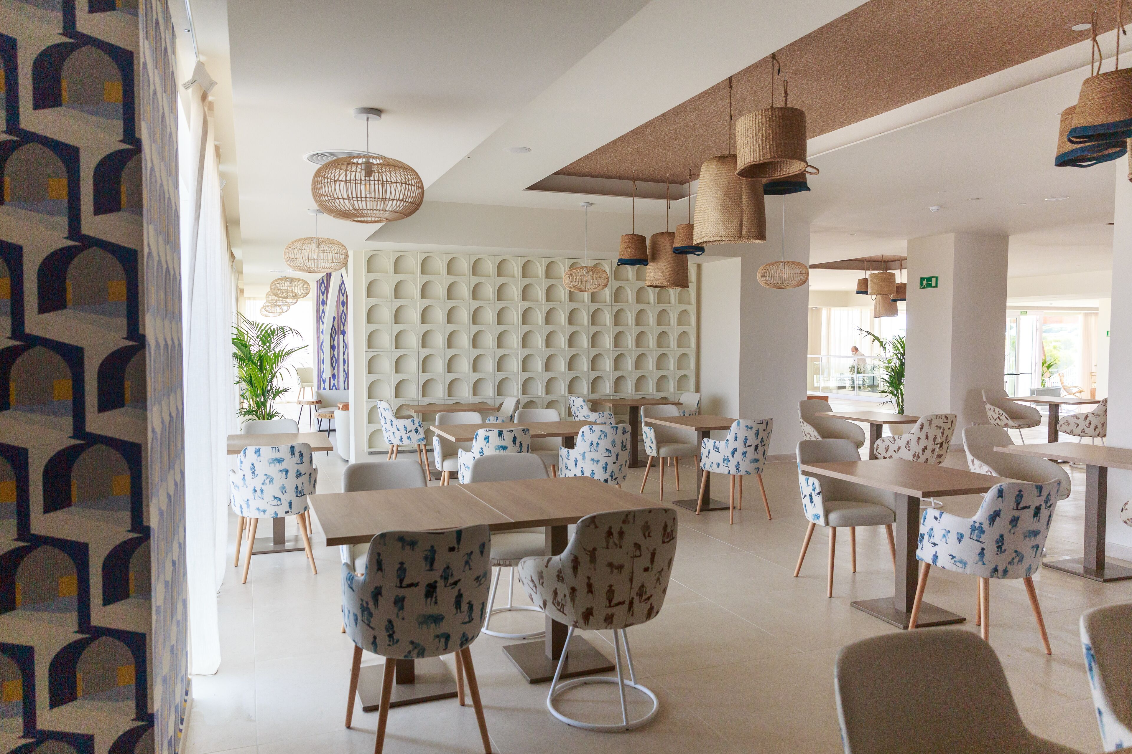 A modern dining area with multiple tables and chairs, a patterned wall, and hanging wicker lamps.