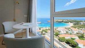 A modern room with a large window offering a view of a coastal town and the sea. - Palladium Hotel Menorca (Mercadal)