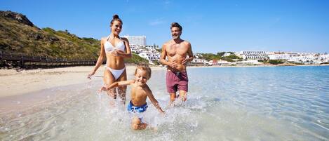 Escena de playa con agua cristalina, una orilla arenosa y edificios al fondo.