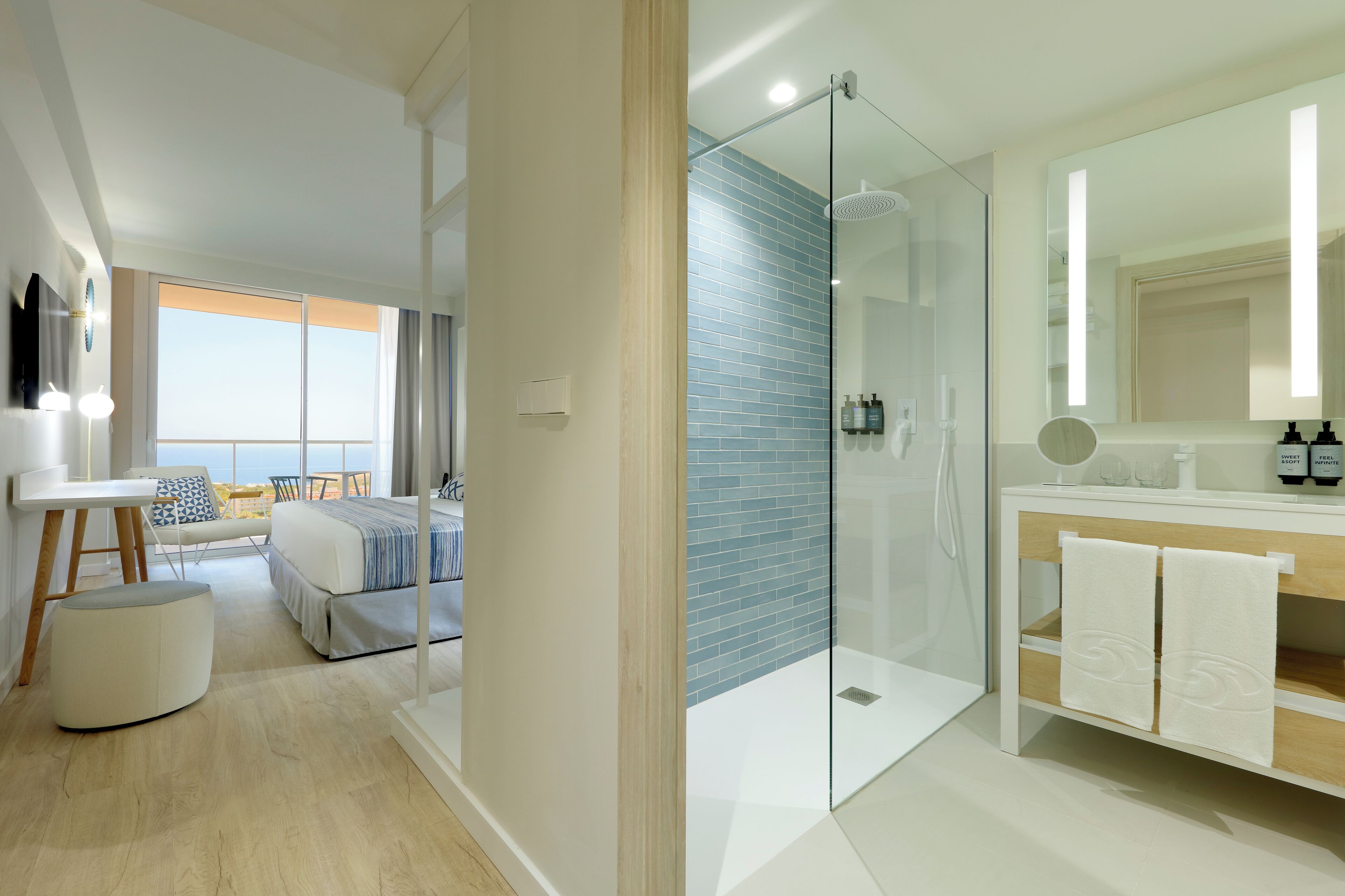 A modern hotel room with a glass-enclosed shower, a large mirror, a wooden vanity, and a balcony with a view.