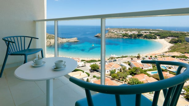 A balcony with a table and chairs overlooking a beach and ocean. - Palladium Hotel Menorca (Mercadal)