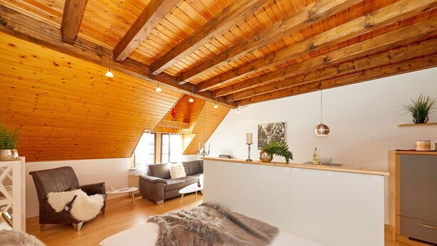 A cozy room with a sloped ceiling, wooden beams, a bed with fluffy pillows, a sofa, a small table, and a kitchenette area. - Altes Eichamt (Bernkastel-Kues)