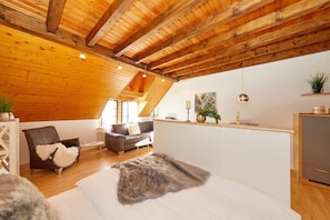 Ein gemütliches Zimmer mit schrägverlaufender Decke, Holzbalken, einem Bett mit weichen Kissen, einer Couch, einem kleinen Tisch und einer Küchenzeile.