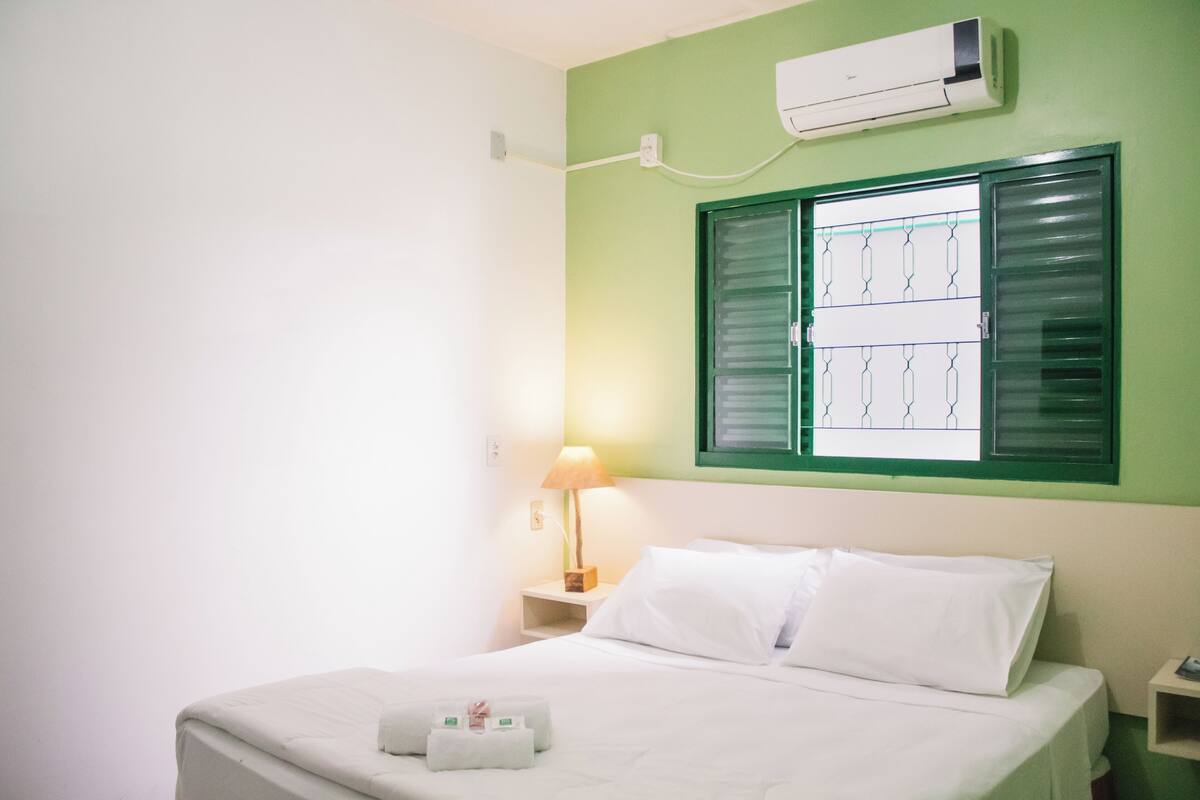 A hotel room with a bed, a nightstand, a lamp, an air conditioner, and a window with green shutters.