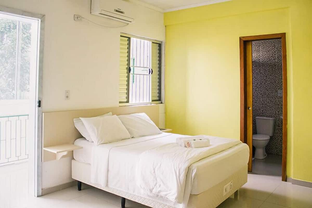 A bedroom with a bed, a bedside table, a window with blinds, and a door leading to a bathroom.