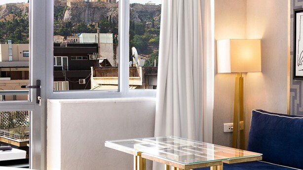Ein modernes Zimmer mit einem Glastisch, zwei Hocker, einem blauen Sofa und Blick auf alte Ruinen durch große Fenster.