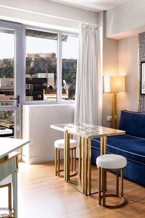 Une pièce moderne avec une table en verre, deux tabourets, un canapé bleu et une vue sur d’anciennes ruines, visible à travers de grandes fenêtres.