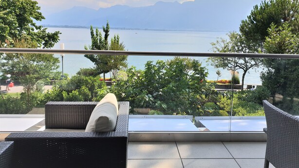 Un balcon doté de meubles en osier, d’une rambarde en verre et offrant une vue sur un lac et des montagnes.
