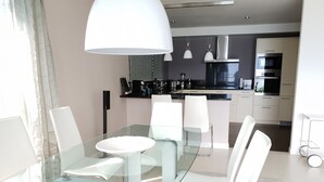 Un espace repas moderne avec une table en verre et des chaises blanches, une cuisine en arrière-plan dotée d’appareils électroménagers en acier inoxydable, et un grand luminaire suspendu blanc au-dessus de la table.