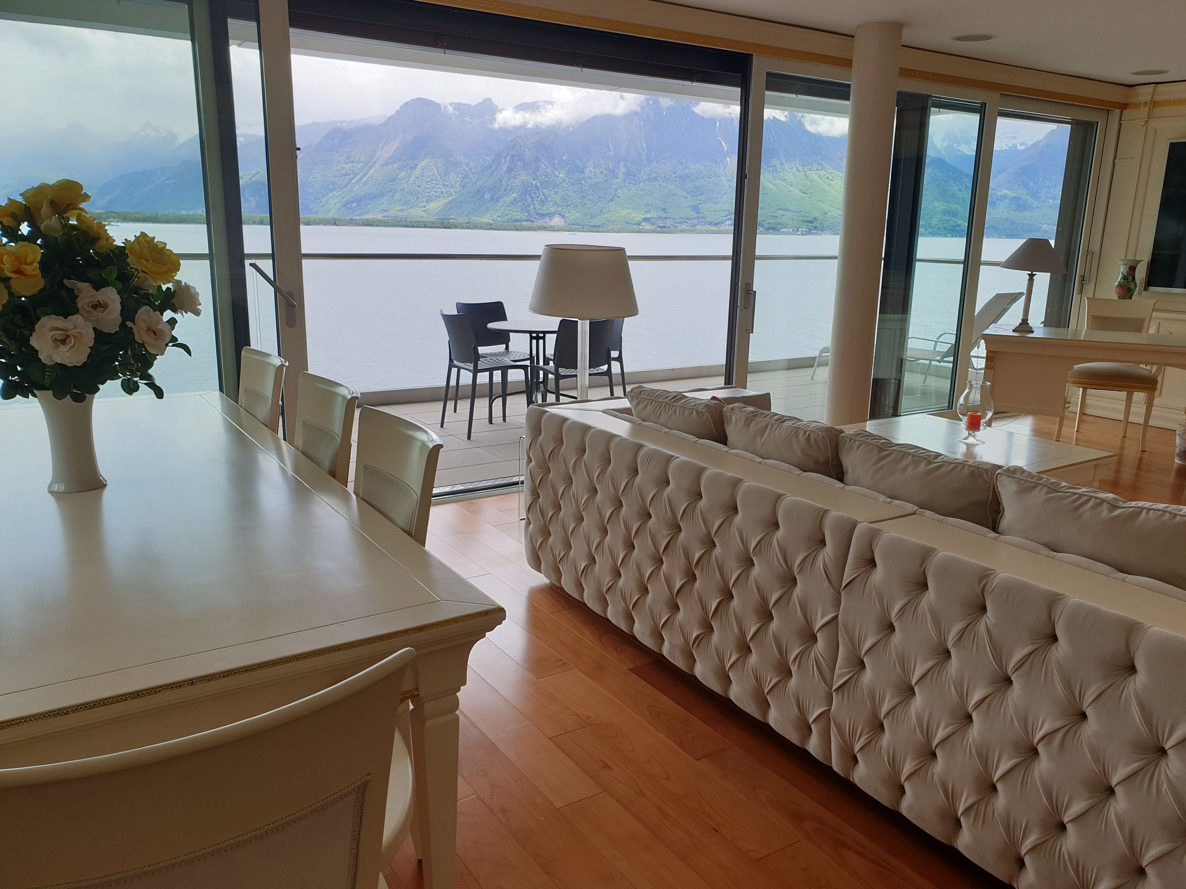 A modern room with a large window offering a mountain view, a plush sofa, a dining table with chairs, and a small table with a lamp and vase.