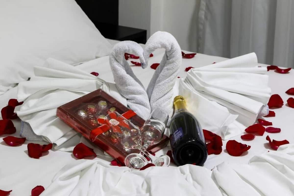 A bed with white linens, a heart-shaped towel arrangement, a champagne bottle, and a tray with glasses and rose petals.