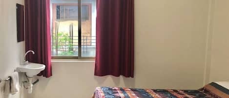 A small, simple room with a bed, a window with curtains, a sink, and an air conditioner.