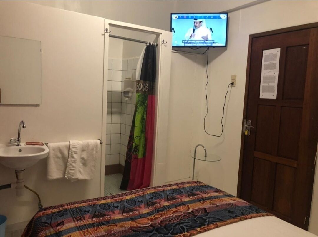 Quarto de hotel com cama, banheiro com chuveiro, pia e televisão fixada na parede.