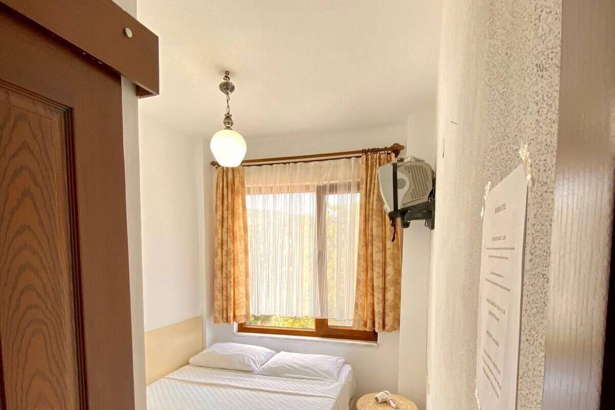 A small, cozy bedroom with a single bed, a bedside table, a window with curtains, and a wall-mounted light fixture.