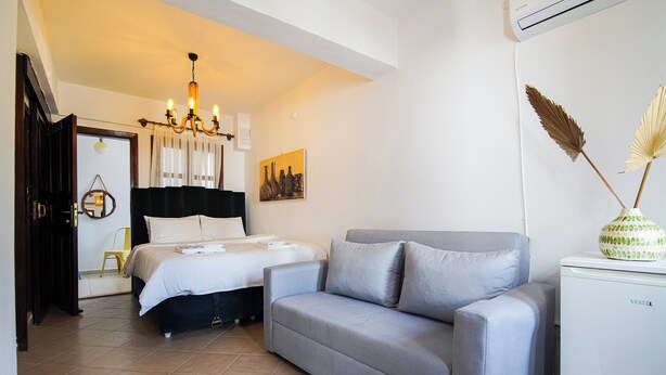 A bedroom with a bed, a grey sofa, a small white fridge, and a chandelier. - Uysal Pansiyon (Bozcaada)