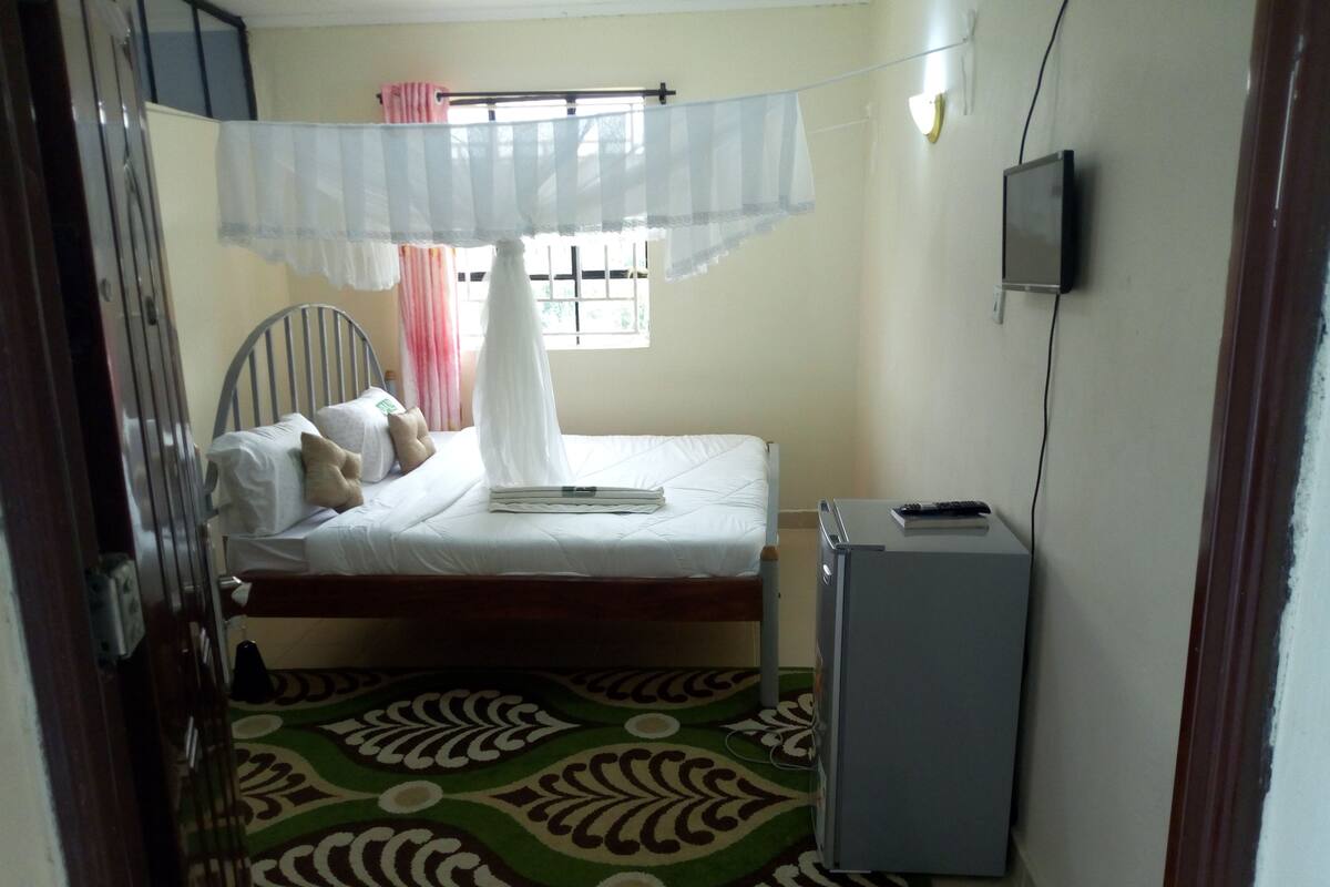 A small, narrow room with a bed, a small desk, a television, and a patterned carpet.