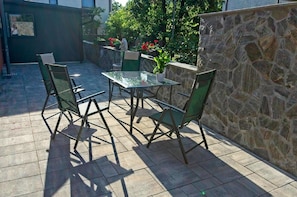 Patio esterno con tavolo in vetro e quattro sedie, circondato da un muro in pietra e piante in vaso.