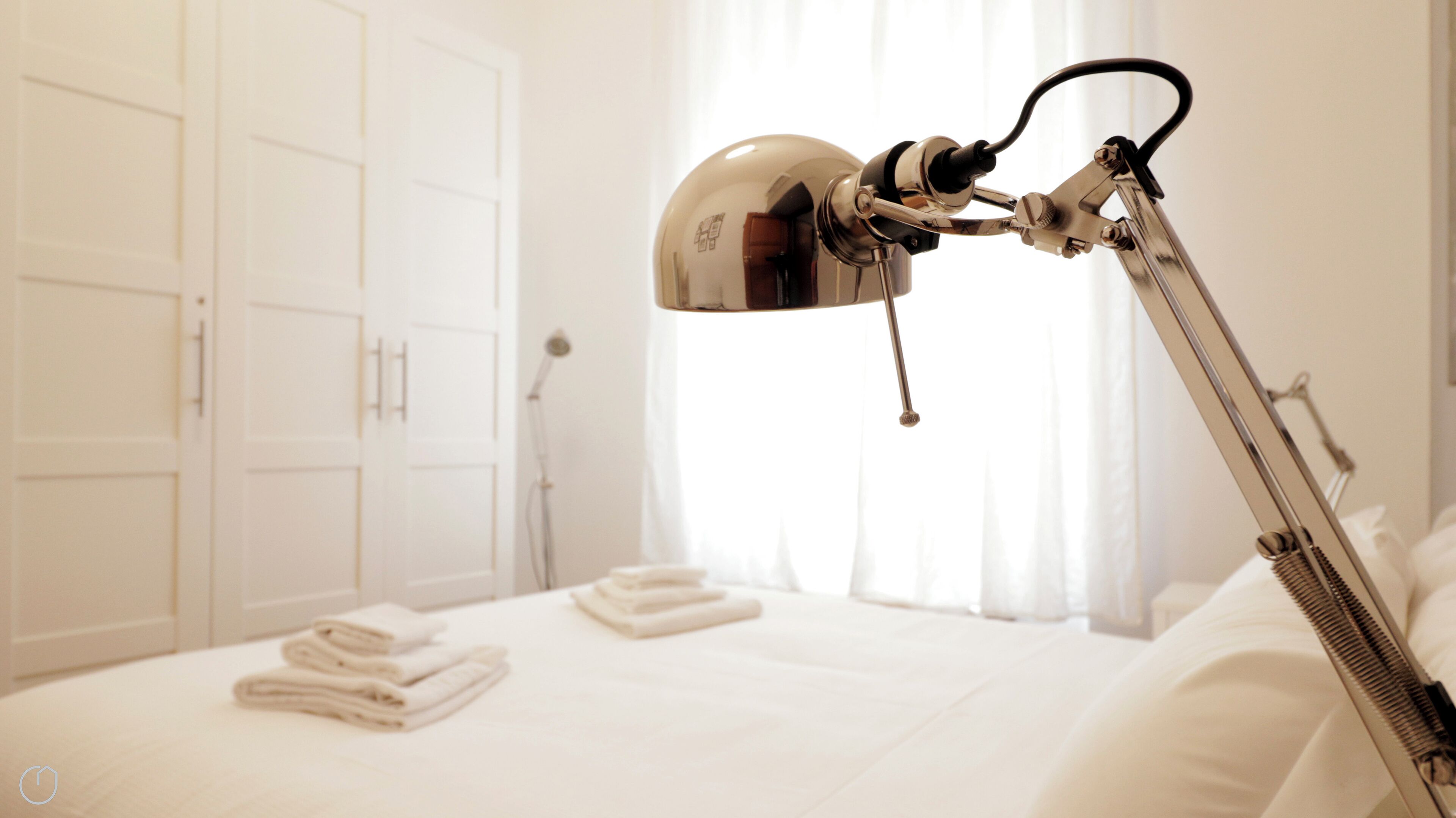 Ein ordentlich bezogenes Bett, gefaltete Handtücher, eine moderne, verstellbare Schreibtischlampe und ein weißer Kleiderschrank in einem Hotelzimmer.