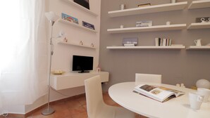 Quarto moderno e bem iluminado, com uma escrivaninha branca, televisão de tela plana e uma mesa de jantar branca com uma revista e xícaras de café. Prateleiras com livros e objetos decorativos são visíveis.