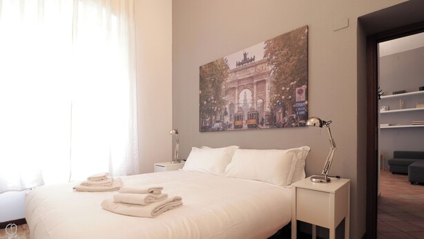 Cama bem arrumada com toalhas dobradas, mesa de cabeceira com abajur e quadro na parede retratando um horizonte urbano.