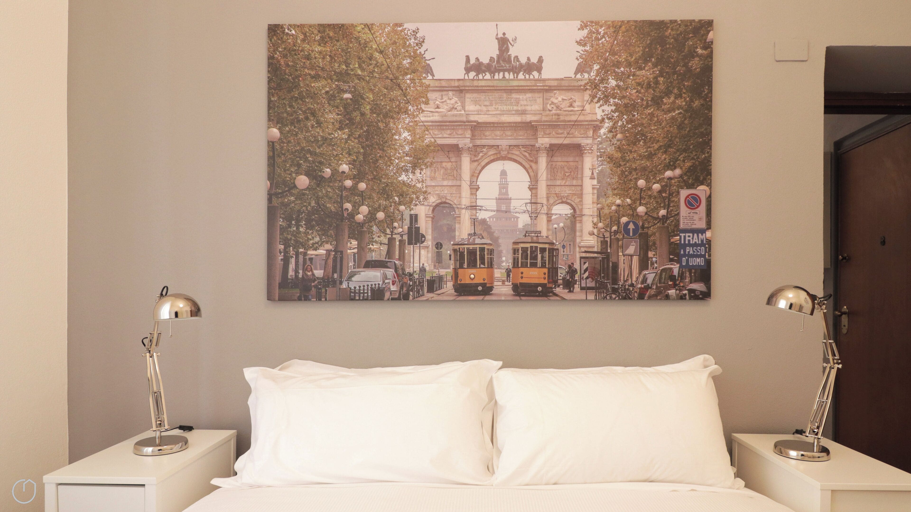 Una habitación de hotel con cama, almohadas blancas, dos mesitas de noche con lámparas y un cuadro grande de un paisaje urbano en la pared.