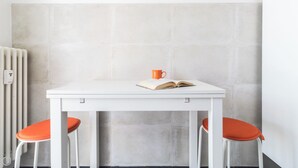 A white table with an open book and an orange mug, flanked by two orange chairs, against a grey tiled wall with a white radiator. - Italianway - Pichi 19 (Milan)