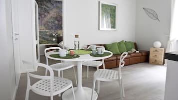 Un salon moderne avec une table ronde blanche, quatre chaises blanches, un canapé en cuir marron agrémenté de coussins verts et une œuvre d’art représentant des feuilles, encadrée, accrochée au mur.