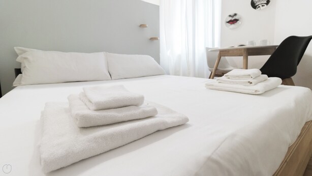 A neatly made bed with folded white towels, a wooden nightstand, and a chair with a black cushion. - Italianway - Pasubio 12 (Milan)