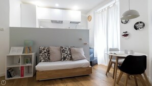 A modern, compact bedroom with a bed, a small desk, and a chair. There is a bookshelf with books and a lamp. The room has a window with curtains and a clock on the wall. - Italianway - Pasubio 12 (Milan)