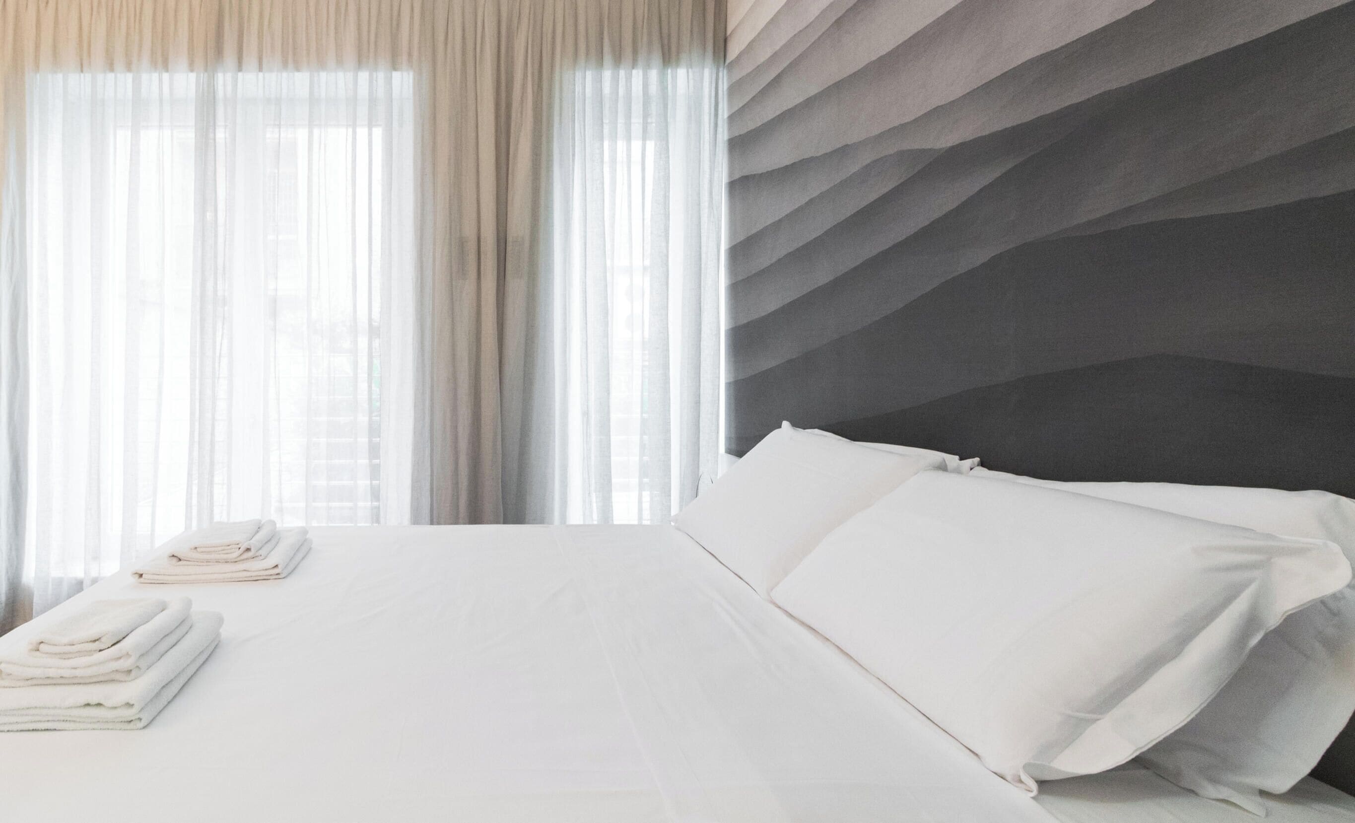 Una habitación de hotel con cama, escritorio y vistas al exterior a través de cortinas translúcidas.