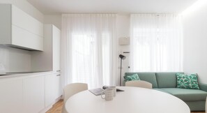 Ein moderner, minimalistischer Wohnraum mit einem runden Esstisch, einer Couch mit gemusterten Kissen und einer Küchenzeile mit weißen Schränken.