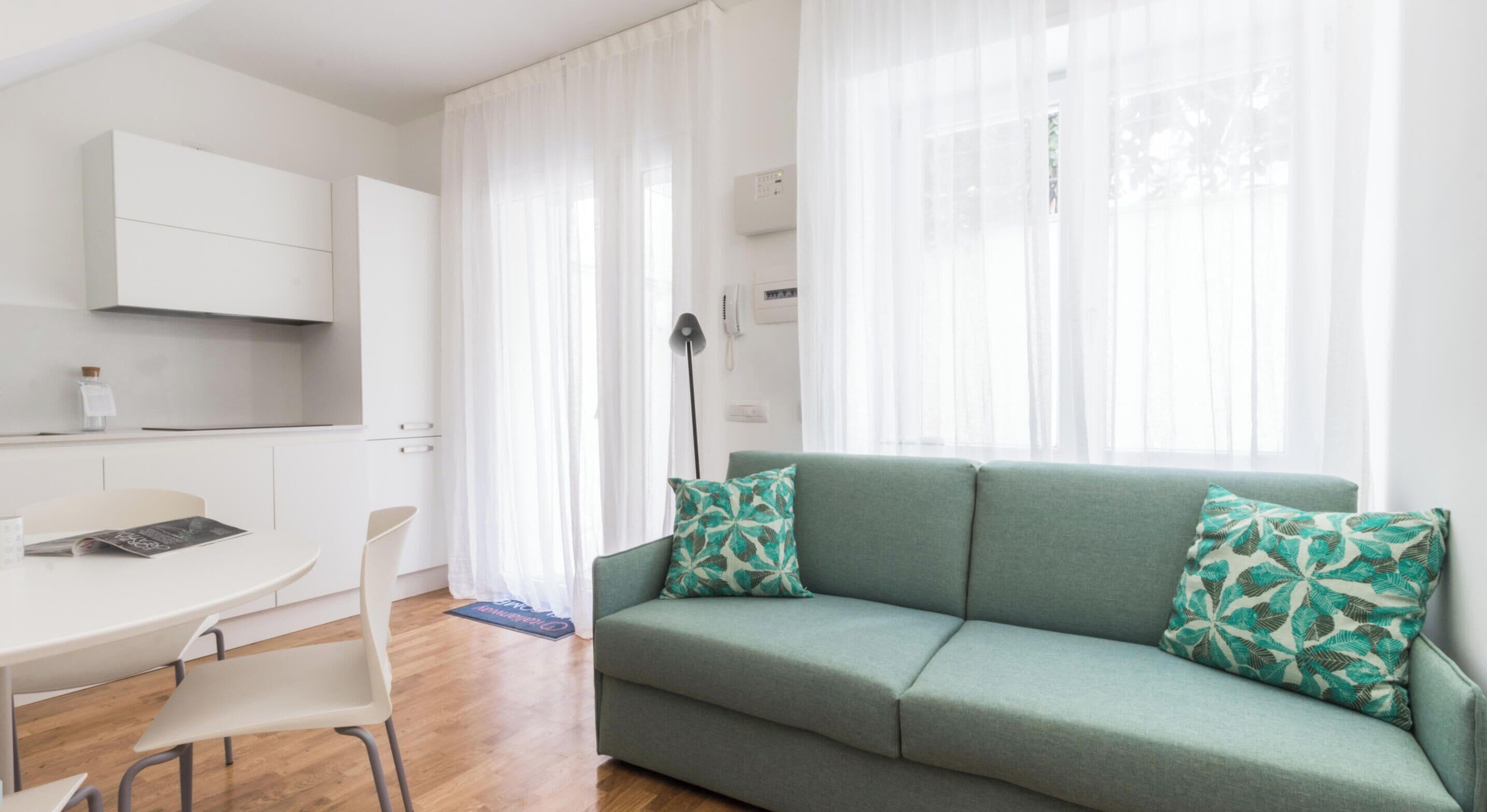 Una sala moderna con un sofá verde, cortinas blancas y una zona de cocina con armarios blancos.