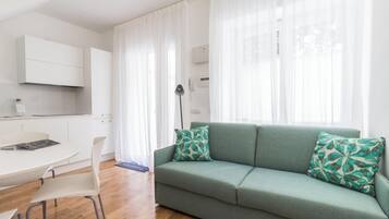 Una sala moderna con un sofá verde, cortinas blancas y una zona de cocina con armarios blancos.