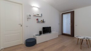 A modern, well-lit room with a flat-screen TV, a small white cabinet, a round ottoman, and a window with sheer curtains. - Italianway Easy - Vigevano 13 (Milan)