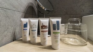Four toiletry items on a wooden surface with a clear glass container and a metal lid in the background. - Italianway Easy - Vigevano 13 (Milan)