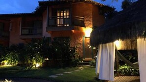 Un edificio de dos pisos con balcón, rodeado de un jardín bien iluminado y una estructura con techo de paja.