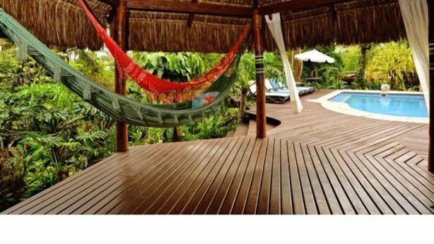 Una terraza de madera con una hamaca, piscina y una estructura con techo de paja.