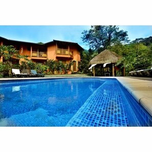 A swimming pool with lounge chairs, a thatched-roof hut, and a two-story building with balconies. - Pousada Forte Rocha (Ilhabela)