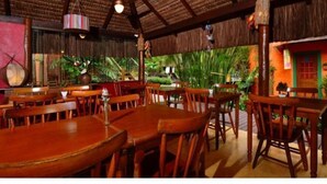 Un restaurante con mesas y sillas de madera, techo de paja y plantas tropicales al fondo.