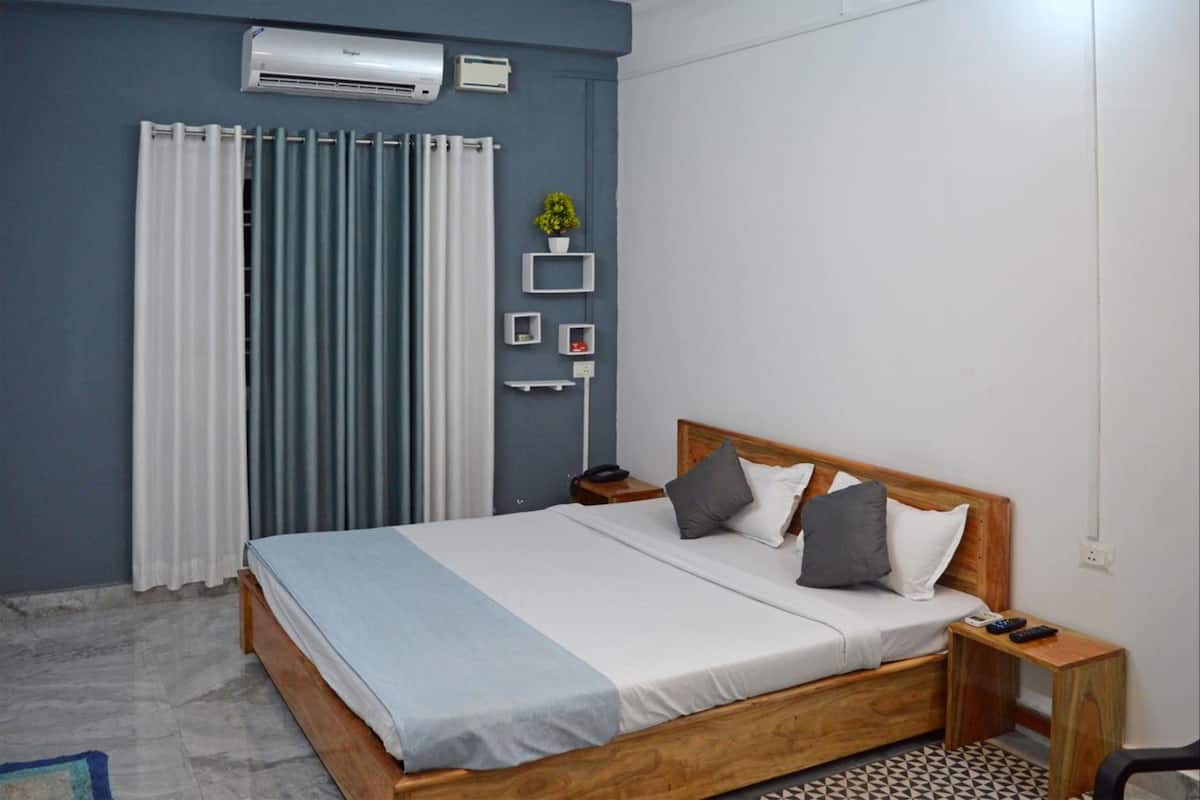 A bedroom with a wooden bed, a nightstand, a wall-mounted air conditioner, a window with curtains, and a small shelf with a plant.