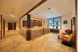 Zona de recepción de un hotel con mostrador de piedra, escritorio de recepción, un reloj y ventana con vistas al exterior.