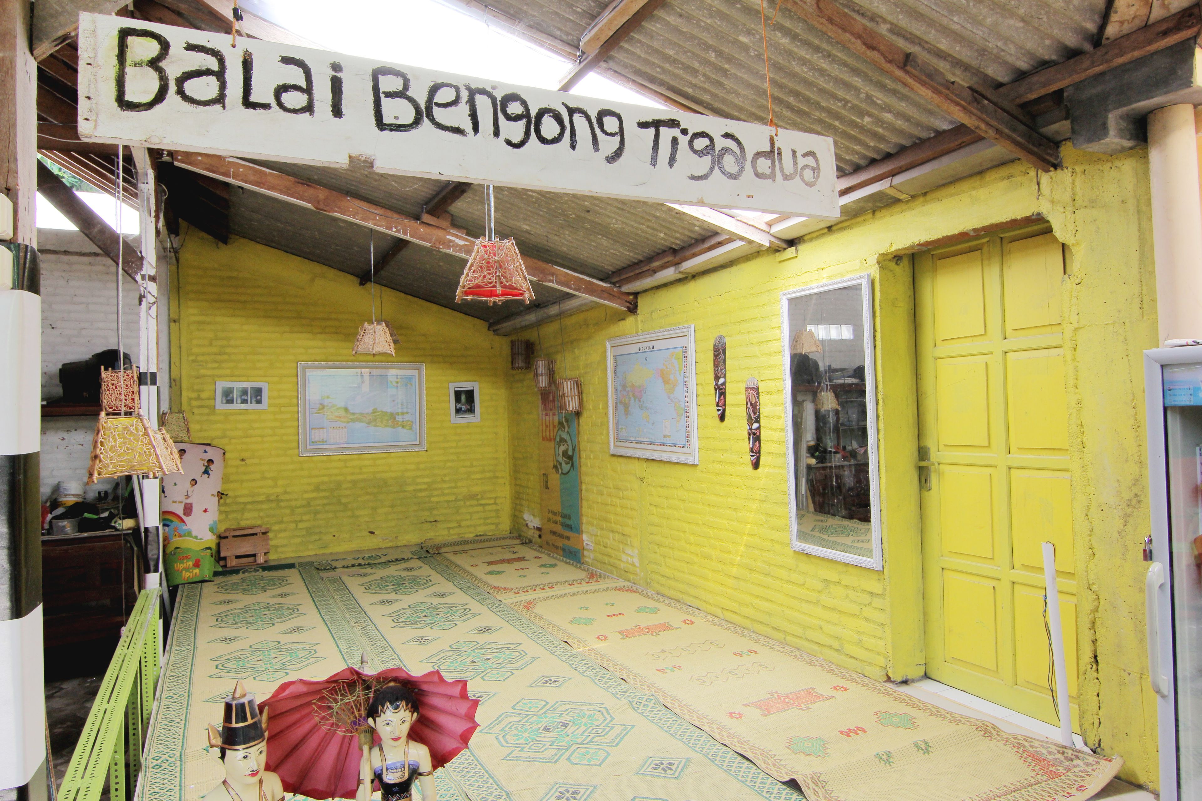 A room with a yellow wall, a wooden door, and a sign reading "Balai Bengong Tigo dua".