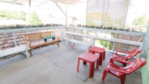 Uma área de estar ao ar livre coberta, com um banco de madeira, cadeiras de plástico vermelhas e uma mesa.