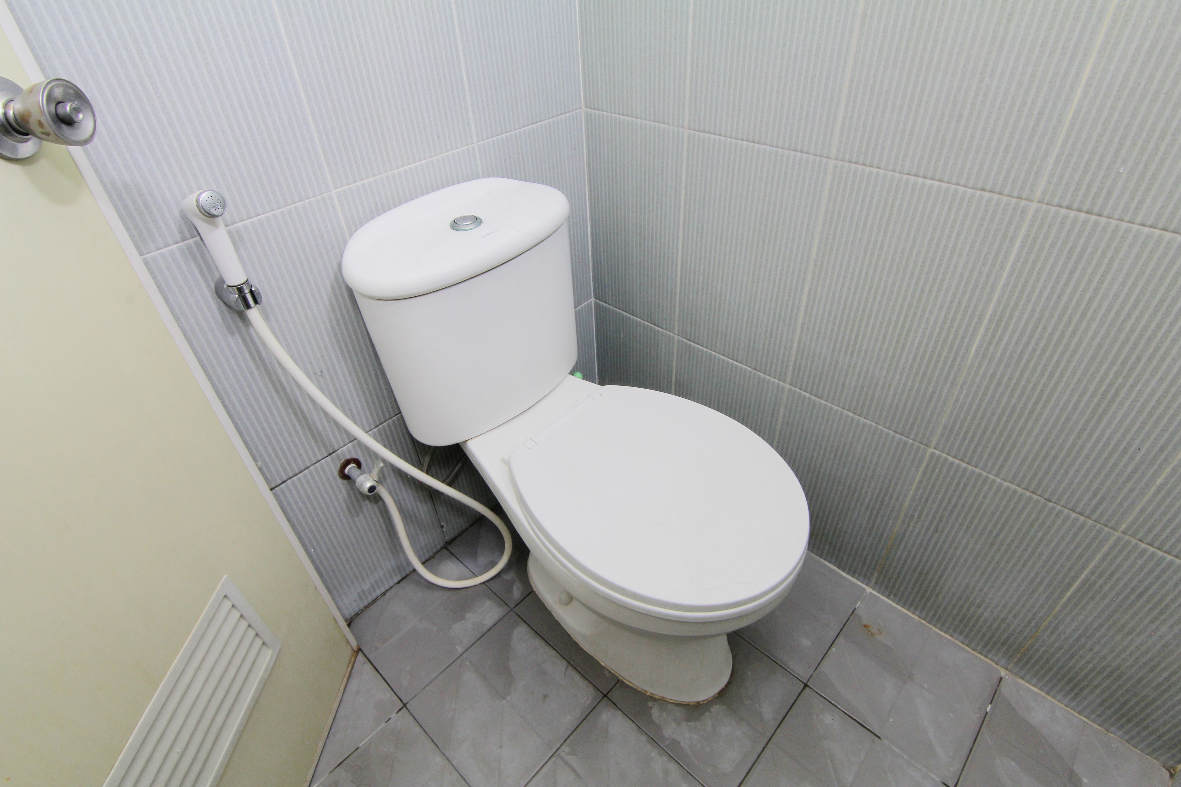 Des toilettes blanches classiques, avec un réservoir et un accessoire de type bidet, dans une salle de bain dont les murs et le sol sont recouverts de carreaux gris.