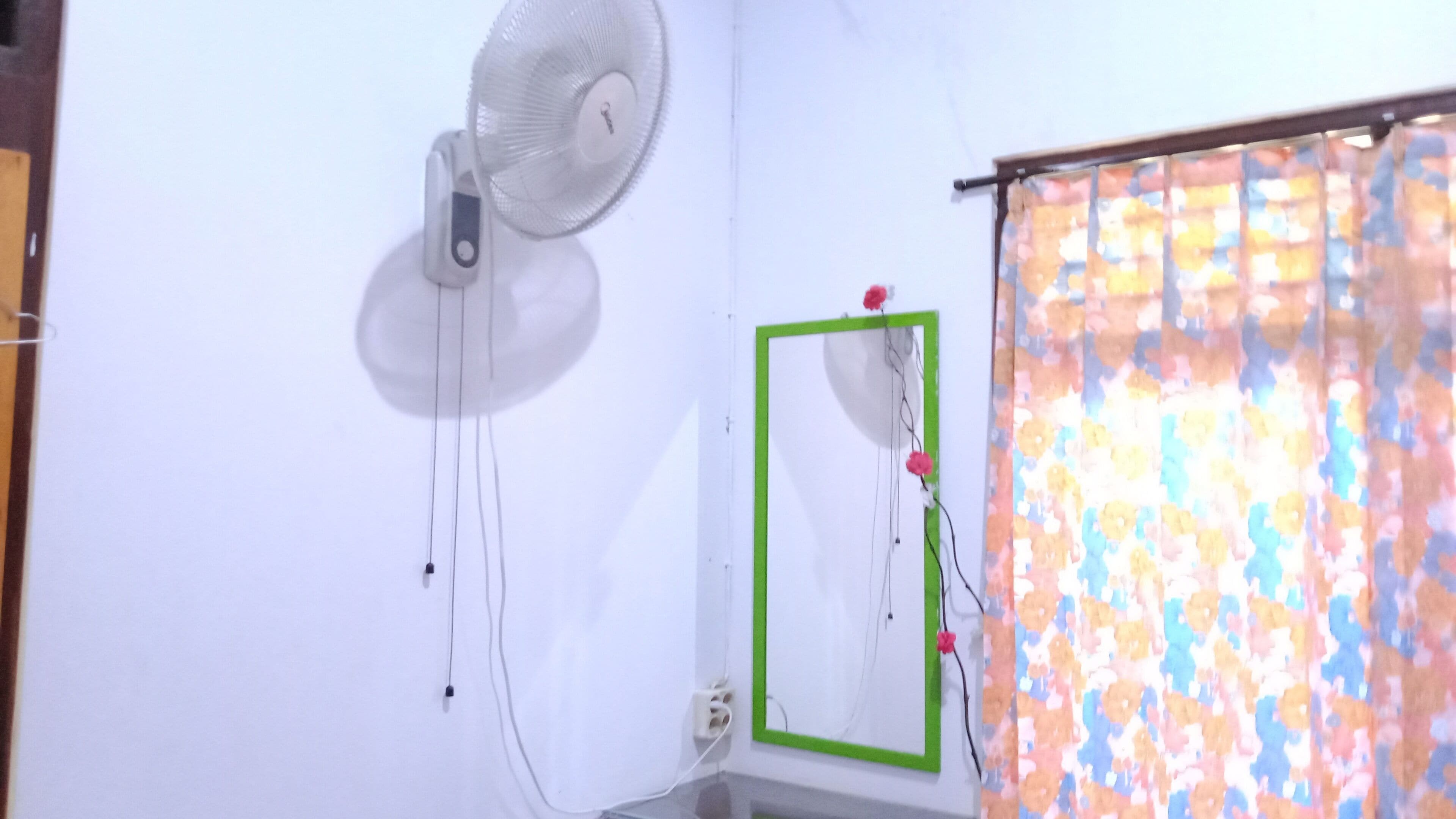 A room with a standing fan, a green-framed mirror, and a window with a floral curtain.