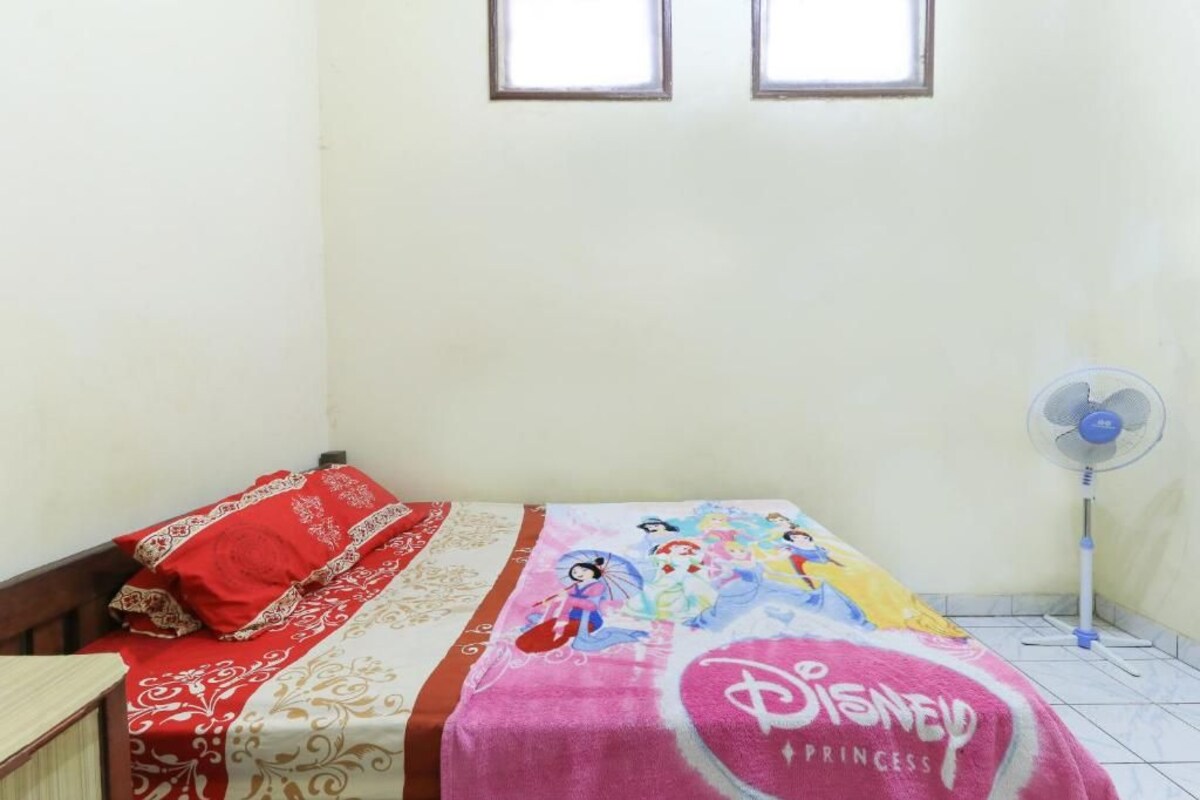A bedroom with a bed, pillows, a Disney Princess blanket, a standing fan, and two windows.